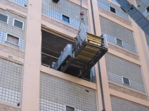 A large industrial machine, expertly handled with rigging services, is being lifted by a crane through a rectangular opening in the side of a multi-story building with glass block windows.