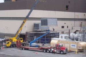 A crane lifts a large industrial unit onto a steel platform beside a commercial building; rigging services crews and trucks are present on site.