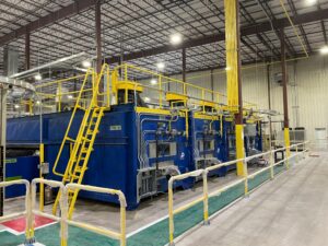Industrial equipment installation featuring blue machines and yellow railings inside a large, well-lit manufacturing facility.