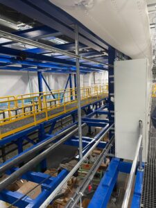 Industrial facility interior in North Andover featuring blue metal framework, yellow safety railings, overhead pipes, and assorted equipment and boxes on the floor—ideal for fabrication installation projects.