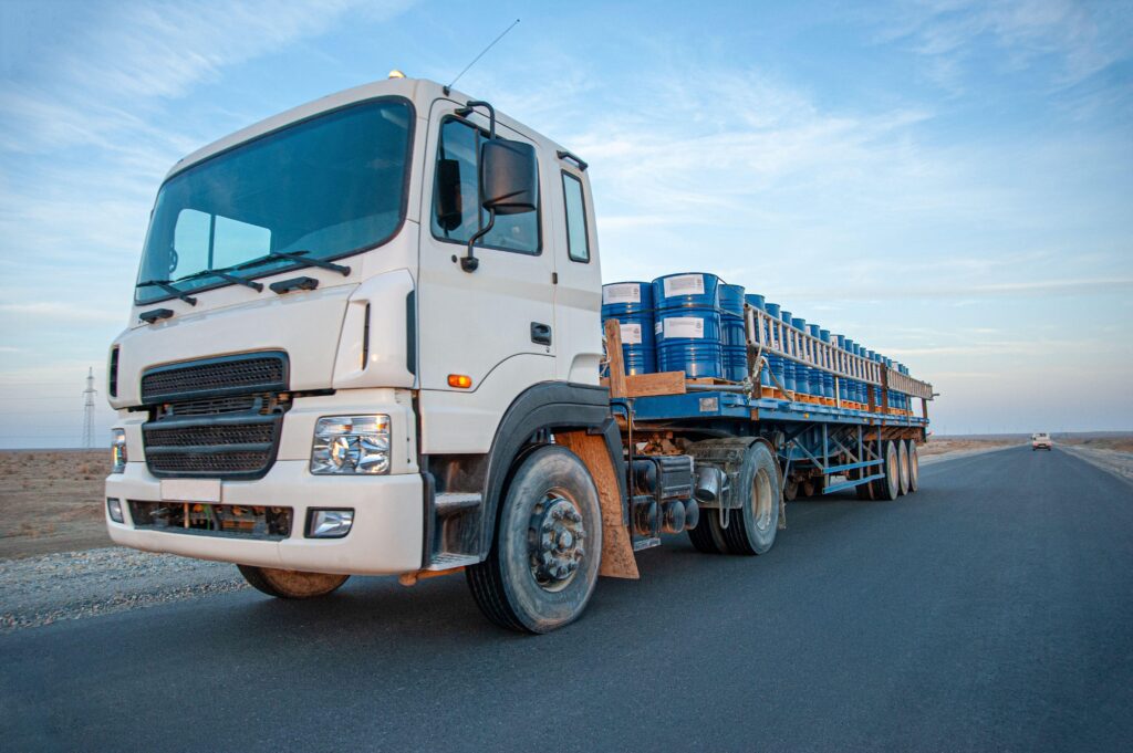 A white semi-truck with a flatbed trailer carrying blue barrels drives on an asphalt road through a dry, open landscape, showcasing efficient transport that can influence rigging services and heavy equipment movers cost.