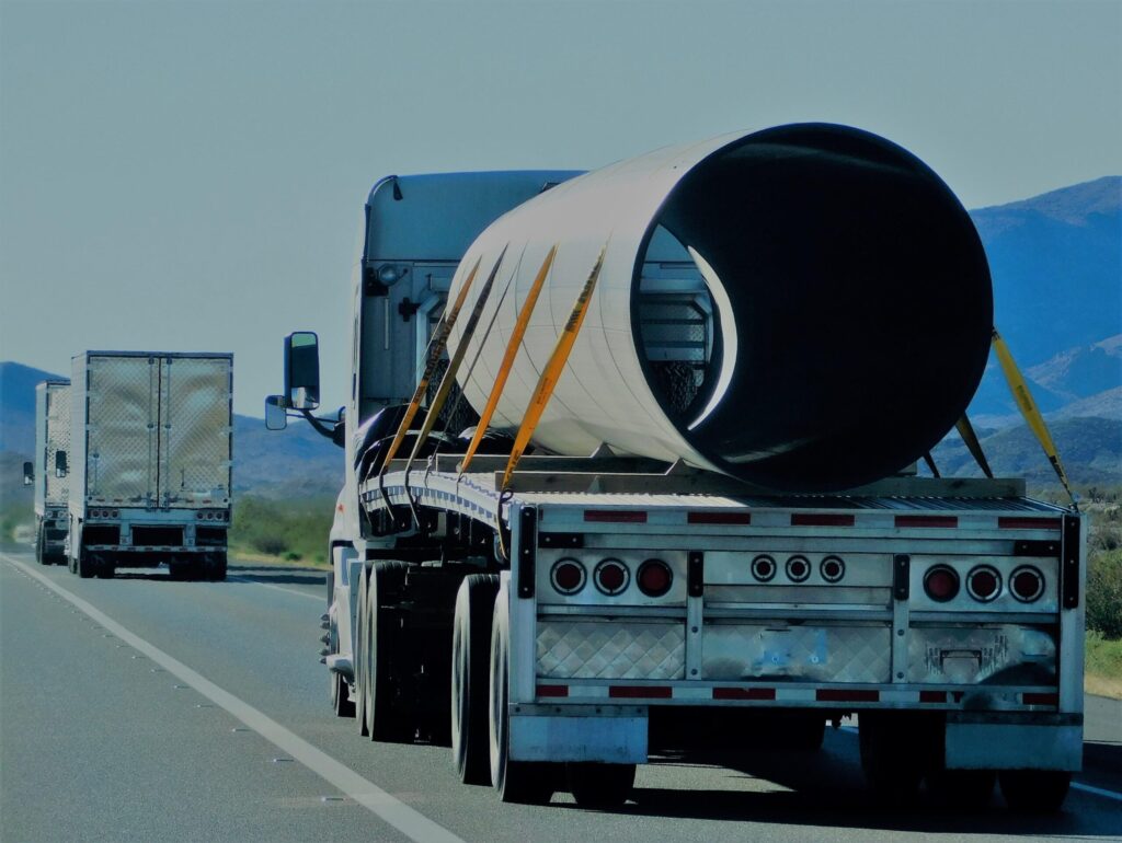 A flatbed truck, used by heavy equipment movers, utilizing professional rigging services, transports a large cylindrical metal pipe secured with straps on a highway, followed by a box truck—highlighting both safety and rigging services cost considerations.
