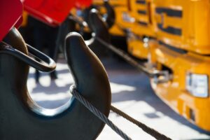 Close-up of a large metal crane hook attached to a steel cable, with yellow heavy equipment movers and construction vehicles visible in the background.