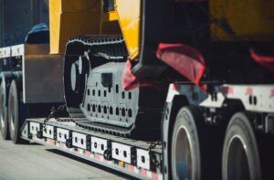 A flatbed truck handles heavy equipment moving, transporting a large tracked construction vehicle with yellow and black parts visible, secured with red straps.