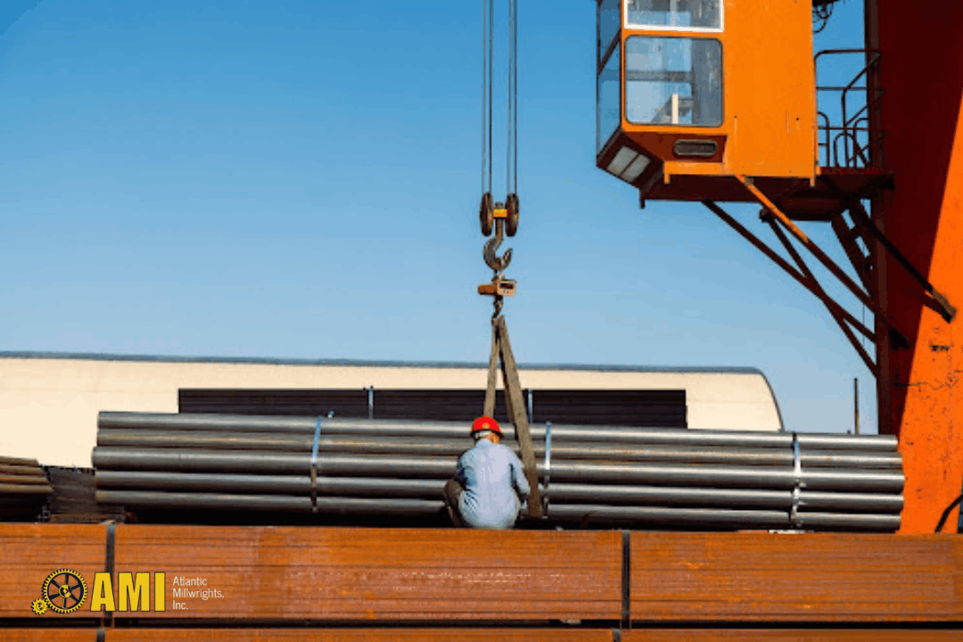 A worker in a red helmet guides a stack of steel pipes being lifted by an orange crane at an industrial site.