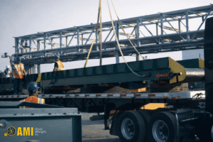 A large metal conveyor is being loaded onto a flatbed truck with a crane at a construction site; workers in safety gear are present.