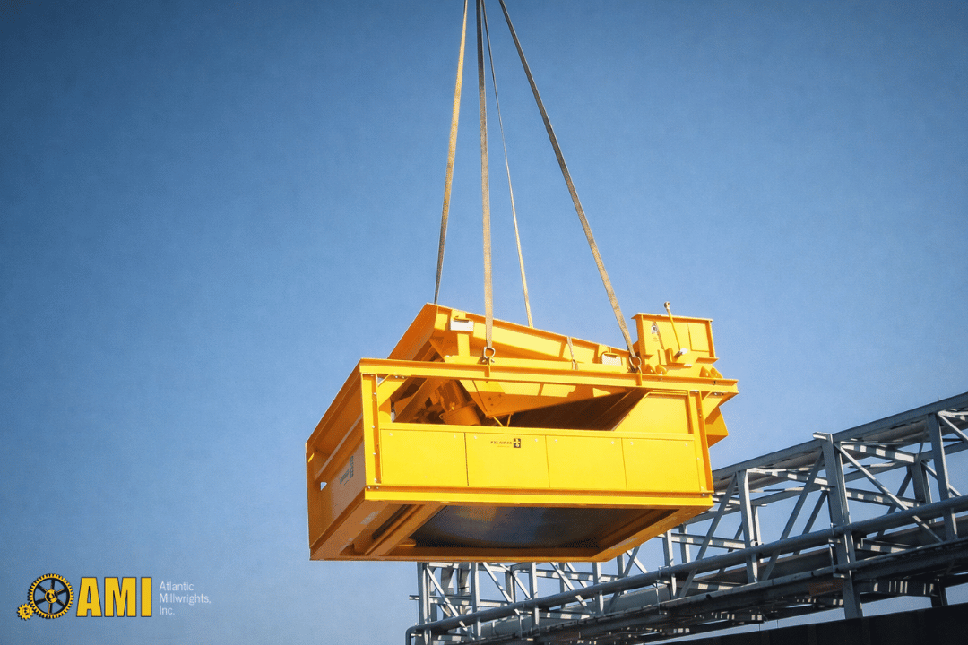 A large yellow industrial machine is being lifted by cables above a metal structure against a clear blue sky. The AMI (Atlantic Minerals, Inc.) logo is visible in the bottom left corner.
