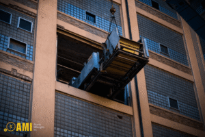 A freight elevator is being lifted by a crane through a large opening in the side of a multi-story industrial building.