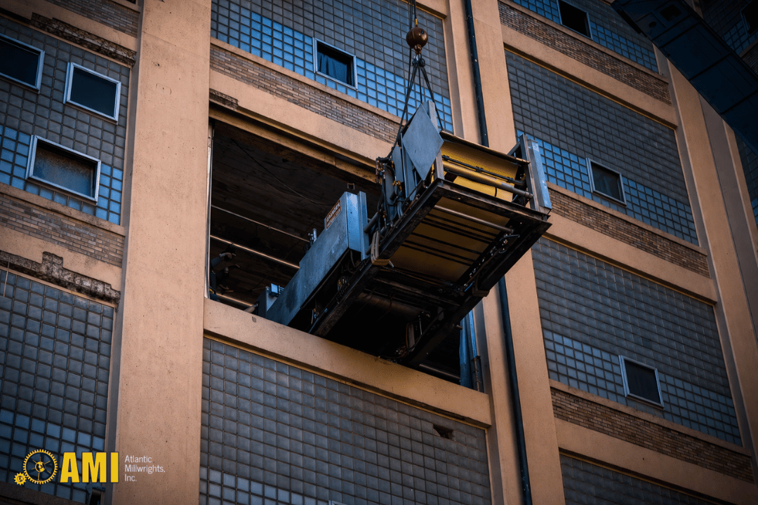 A freight elevator is being lifted by a crane through a large opening in the side of a multi-story industrial building.