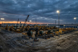A fenced lot serves as a heavy equipment storage area, containing numerous construction vehicles and equipment, including excavators and cranes, under bright lights at dusk with a cloudy sky.